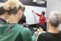 Eine Vortragende zeigt mit ihrer Hand auf einen Text auf einem Whiteboard während einige Teilnehmerinnen und Teilnehmer ihr zuhören.