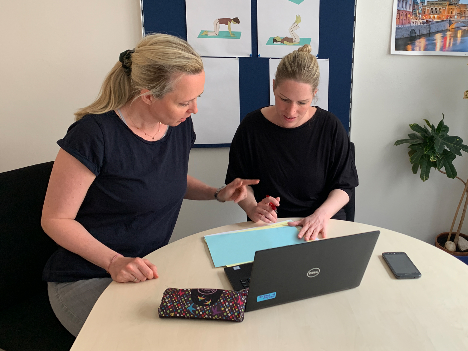 Foto von zwei Frauen welche an einem Schreibtisch mit darauf befindlichem Laptop sitzen und auf ein Dokument blicken.